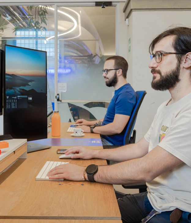 Due colleghi concentrati sul loro lavoro in uno spazio di ufficio condiviso presso Nana Bianca, Firenze. L'ambiente di coworking moderno include monitor doppi e postazioni ergonomiche, favorendo la produttività in un contesto collaborativo.